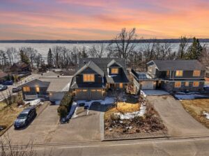Aerial view of a lakeside neighborhood at sunset featuring a beautiful detached home in Shanty Bay, with snow patches and bare trees along the shore.